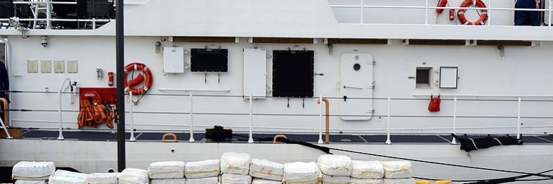 Seized bales of cocaine are stacked in front of a ship.