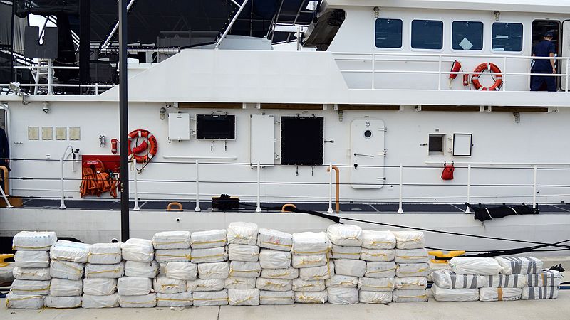 Seized bales of cocaine are stacked in front of a ship.