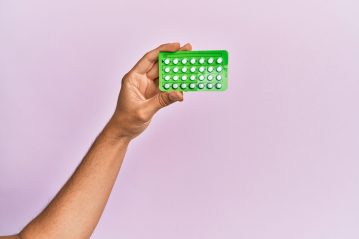 A young person's hand holding birth control pills with a pink background