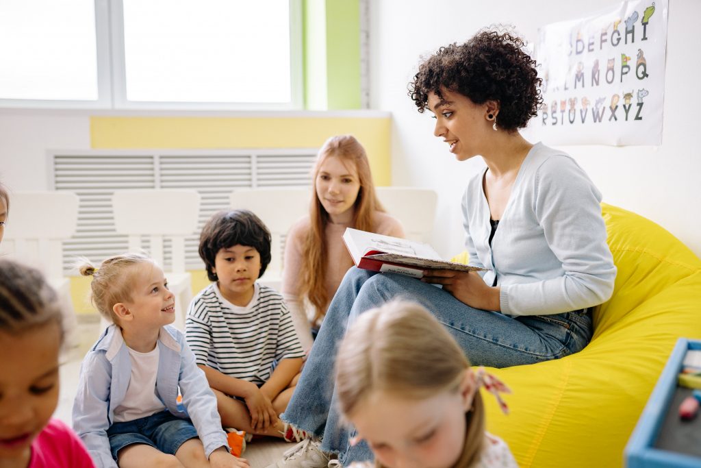 A female teacher sits in a beanbag in a well-lit classroom. She is reading aloud to multiple young children.