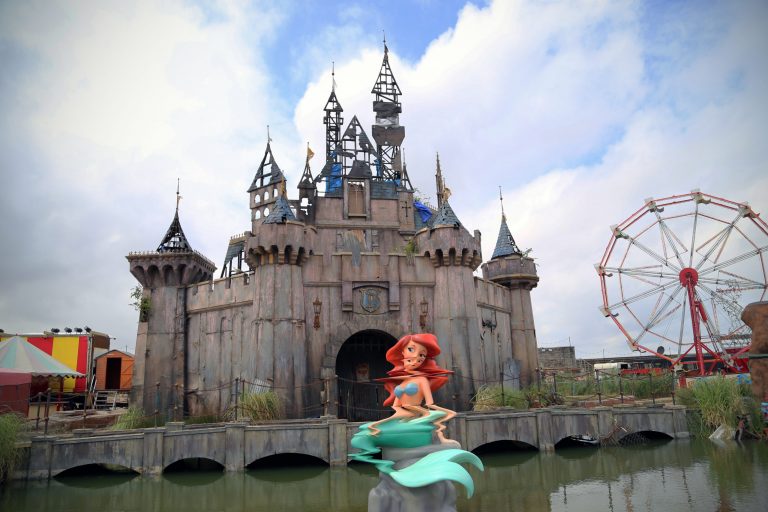 Banksy Dismaland featuring a dilapidated fairytale castle with a warped and glitched statue of a mermaid in front