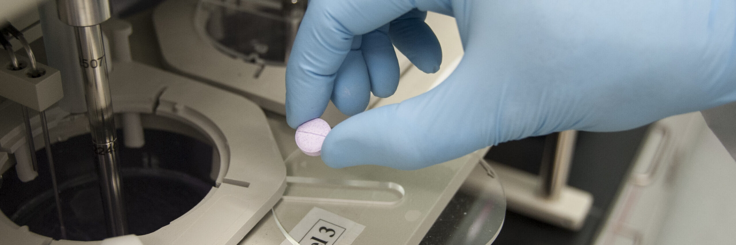 An image of a pill being held by a blue nitrile glove. machines for drug research and pill testing are visible. Image Source: Michael J. Ermarth, via: Wikimedia Commons