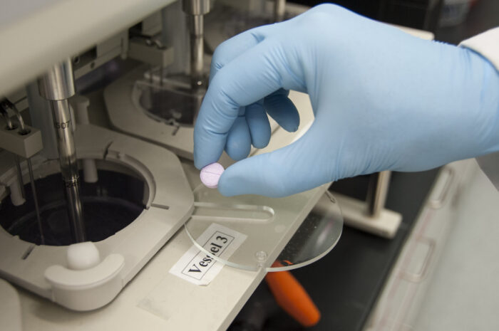 An image of a pill being held by a blue nitrile glove. machines for drug research and pill testing are visible. Image Source: Michael J. Ermarth, via: Wikimedia Commons