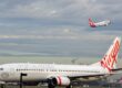 An image of a Virgin Airlines plane on the ground at an airport, while a Qantas Airlines plane is visible in the sky. Image source: Simon_sees, via Wikimedia Commons