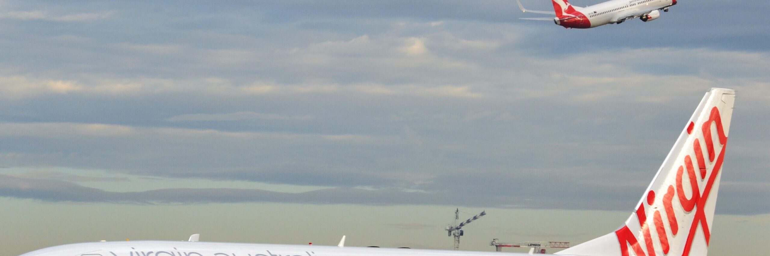 An image of a Virgin Airlines plane on the ground at an airport, while a Qantas Airlines plane is visible in the sky. Image source: Simon_sees, via Wikimedia Commons