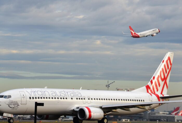 An image of a Virgin Airlines plane on the ground at an airport, while a Qantas Airlines plane is visible in the sky. Image source: Simon_sees, via Wikimedia Commons