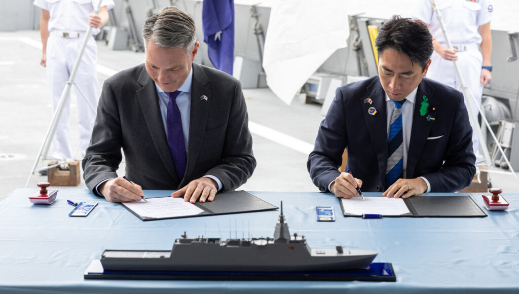 Richard Marles and Koizumi Shinjiro sign the deal in Melbourne with a model of the Mitsubishi Mogami frigate in the foreground. Photo: Department of Defence.
