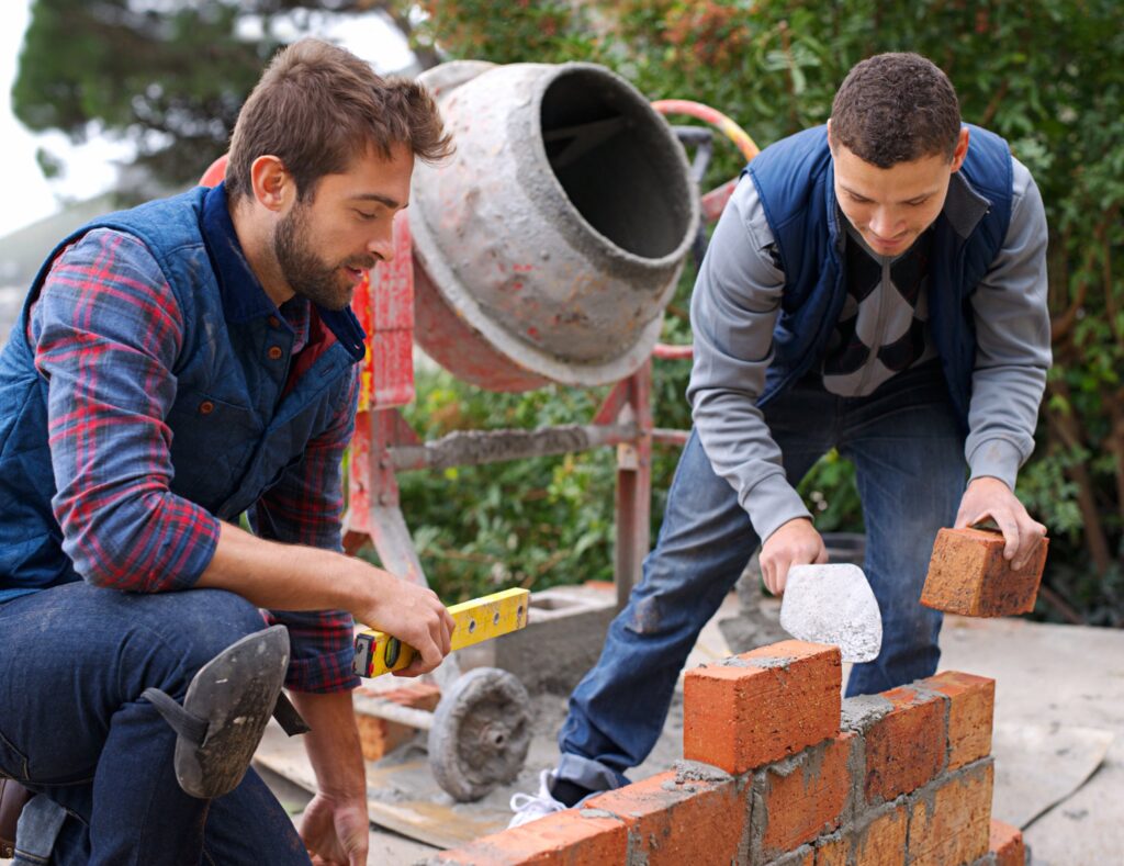 Two tradies laying a brick wall beside a cement mixer, illustrating building apprentices on a housing construction site